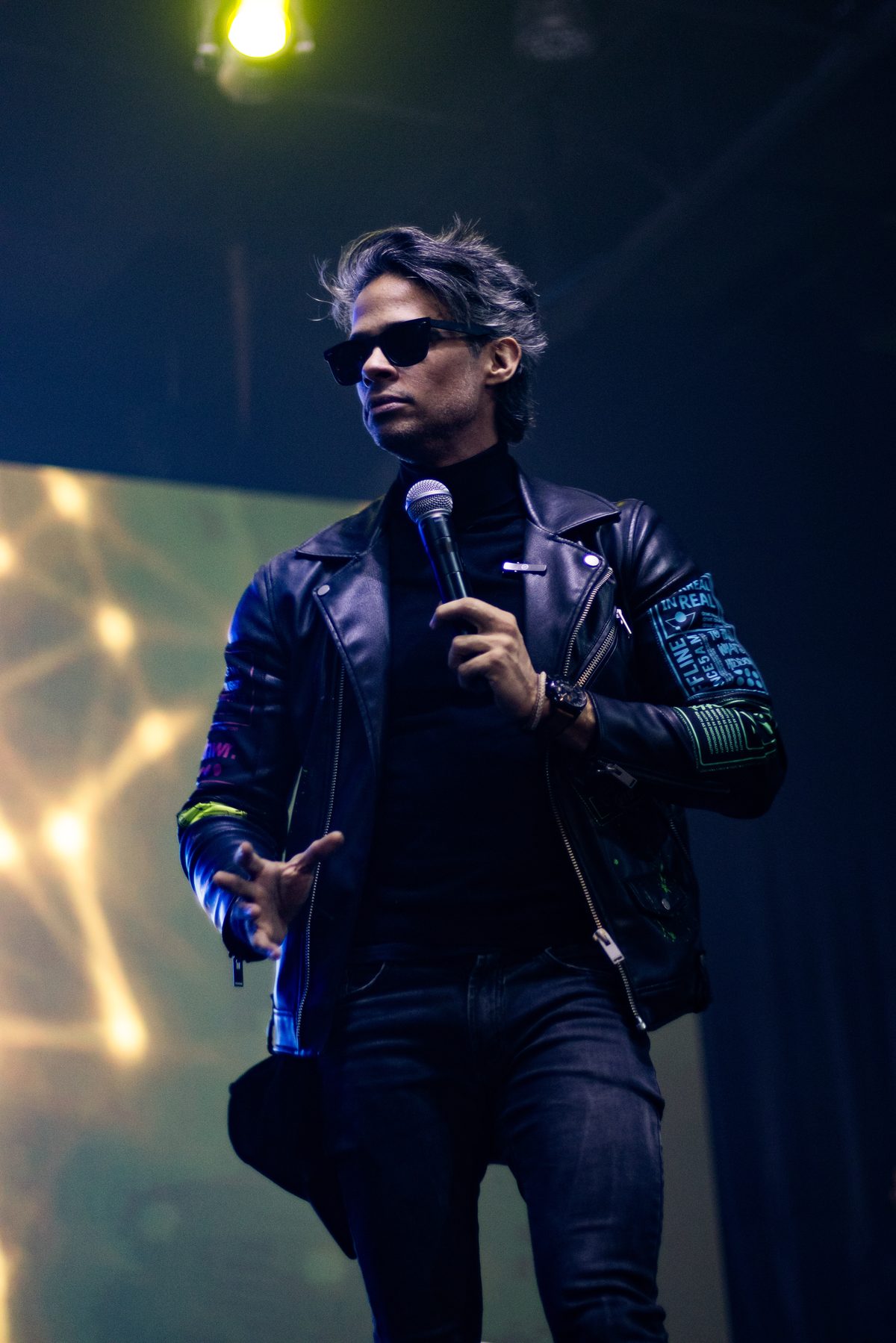 Marcos Strider no palco, de jaqueta de couro e óculos escuros, com o microfone na mão
